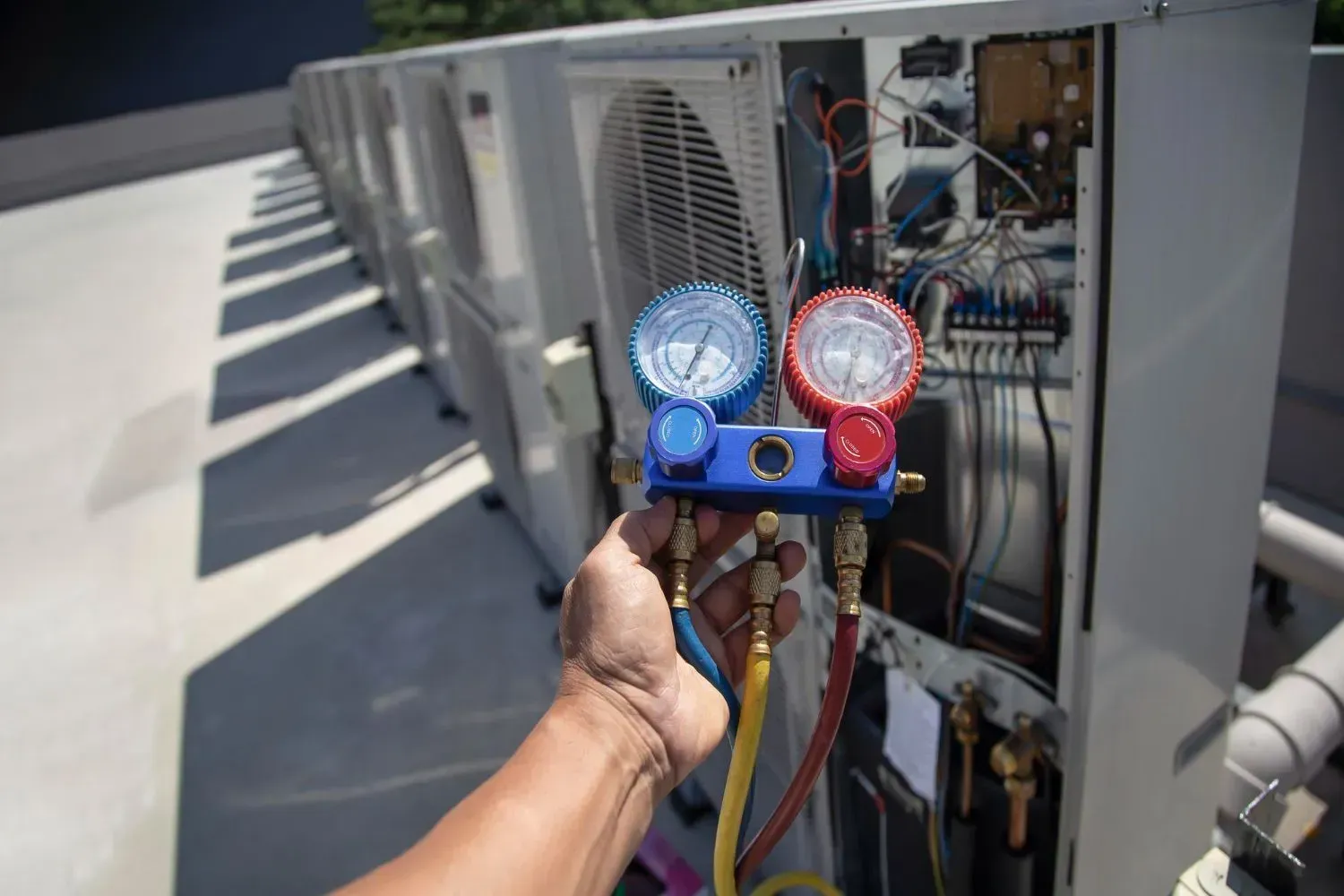 Hand holding HVAC manifold gauges beside an open rooftop air-conditioning unit
