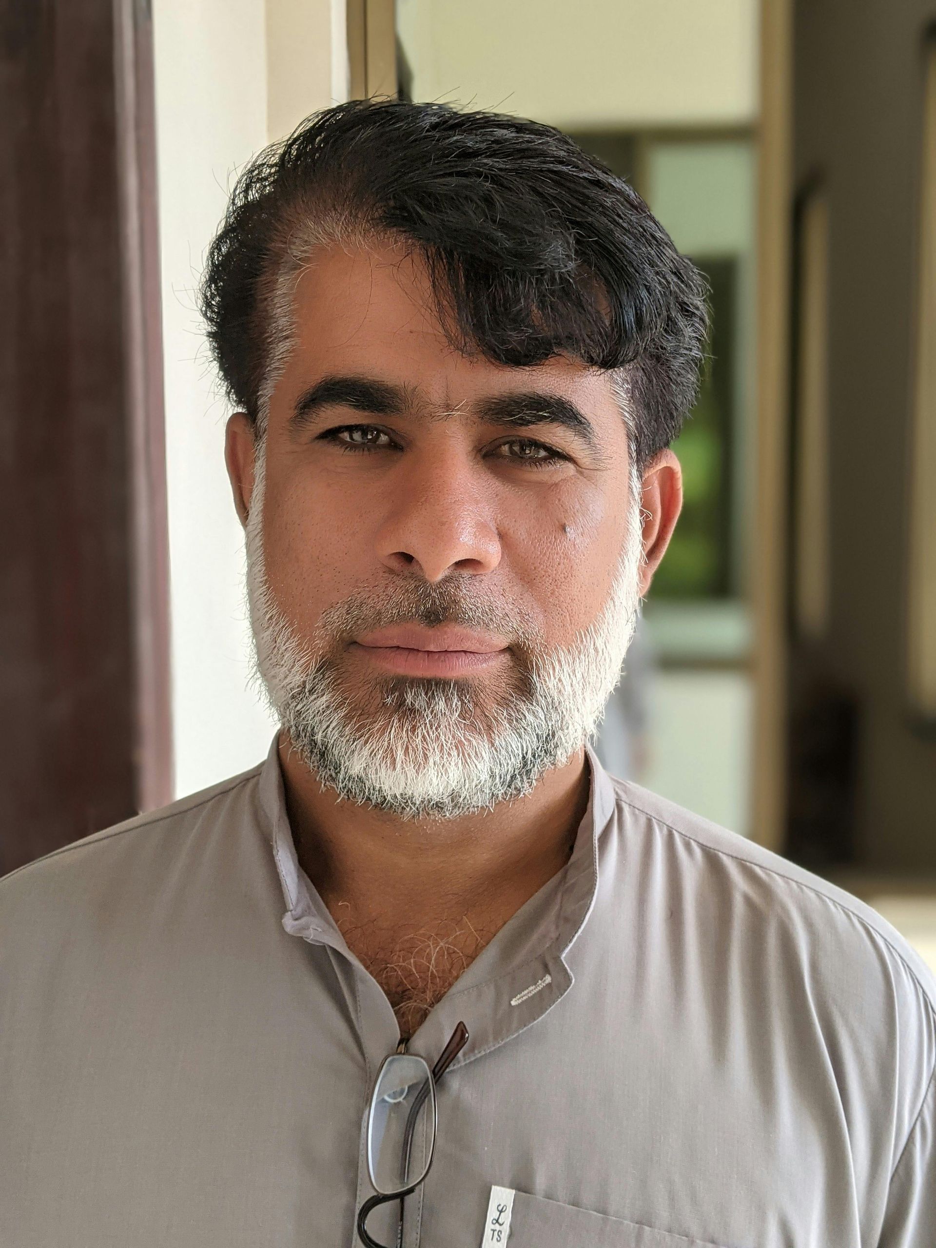 Man standing indoors in a beige shirt, facing the camera with a neutral expression.