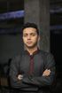 Person in a dark traditional shirt stands with arms crossed against a blurred indoor background.