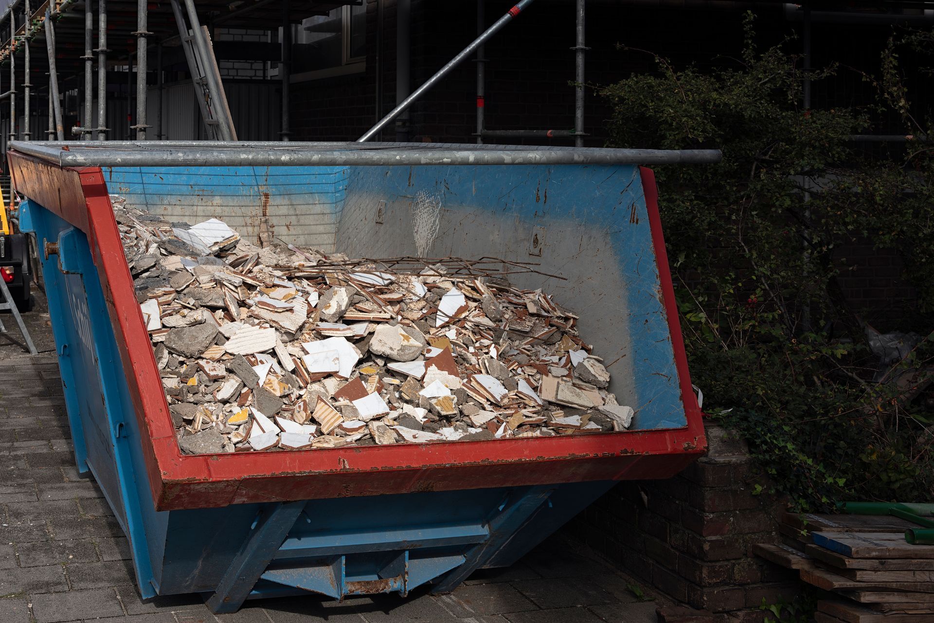 A blue and red skip filled with construction debris sits beside a building with scaffolding.