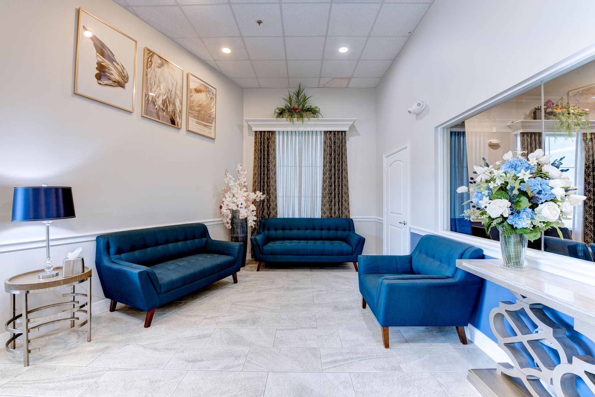 Long hallway with framed artwork, patterned chairs, side tables, and lamps in a softly lit waiting area.