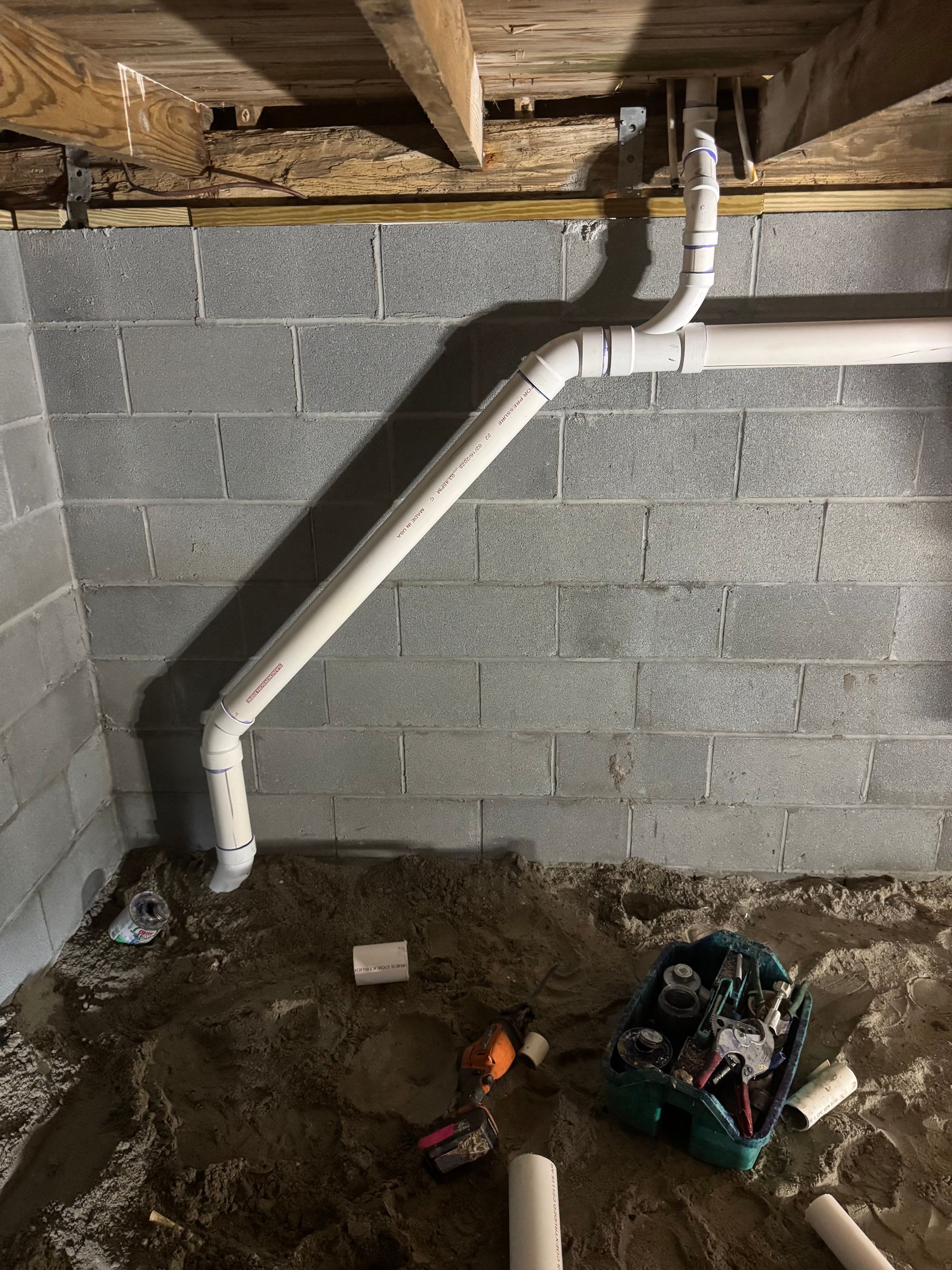 White PVC pipes installed against a concrete block basement wall. Plumbing tools and debris are on the floor.