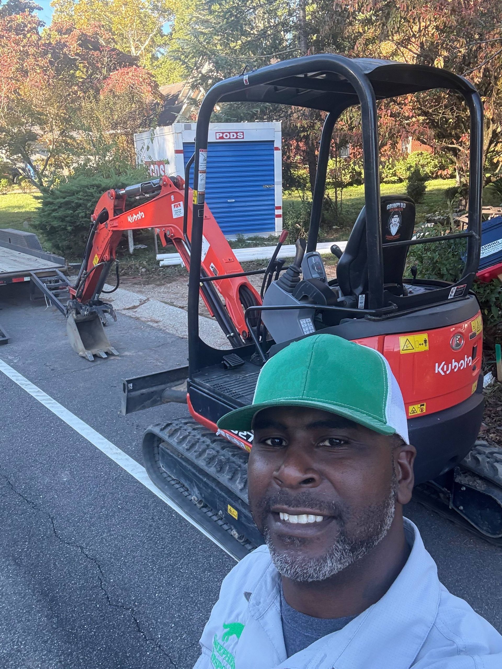 Man in green cap smiles, standing by orange Kubota excavator.