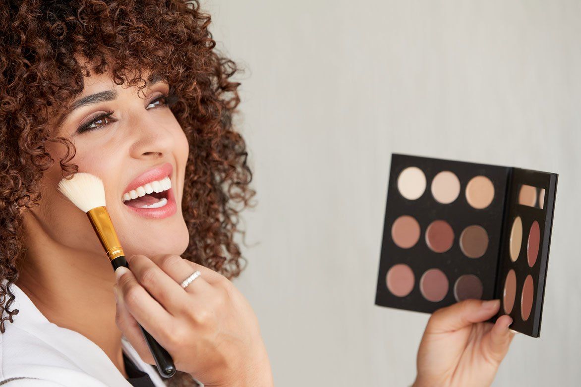 A woman is smiling while applying RoryLee® Makeup to her face.