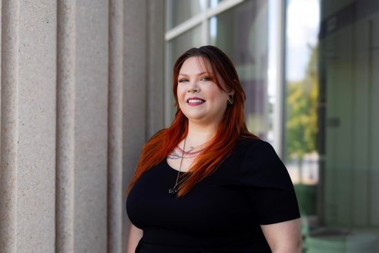 About Me Woman with red-toned hair in black top smiles, standing beside a stone building with glass windows, outdoors.