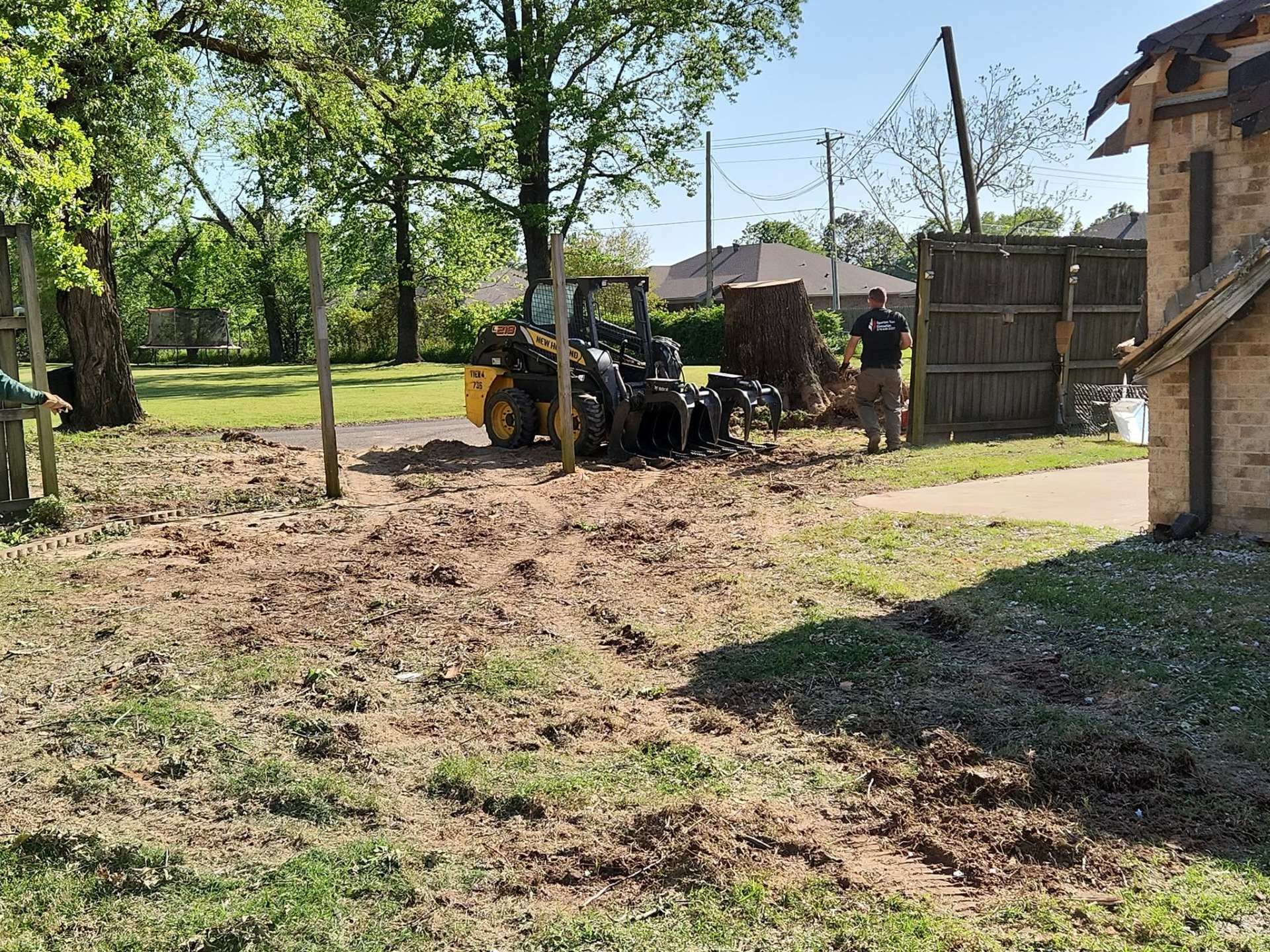 A tractor clears mud in a yard beside trees and a house on a sunny day.