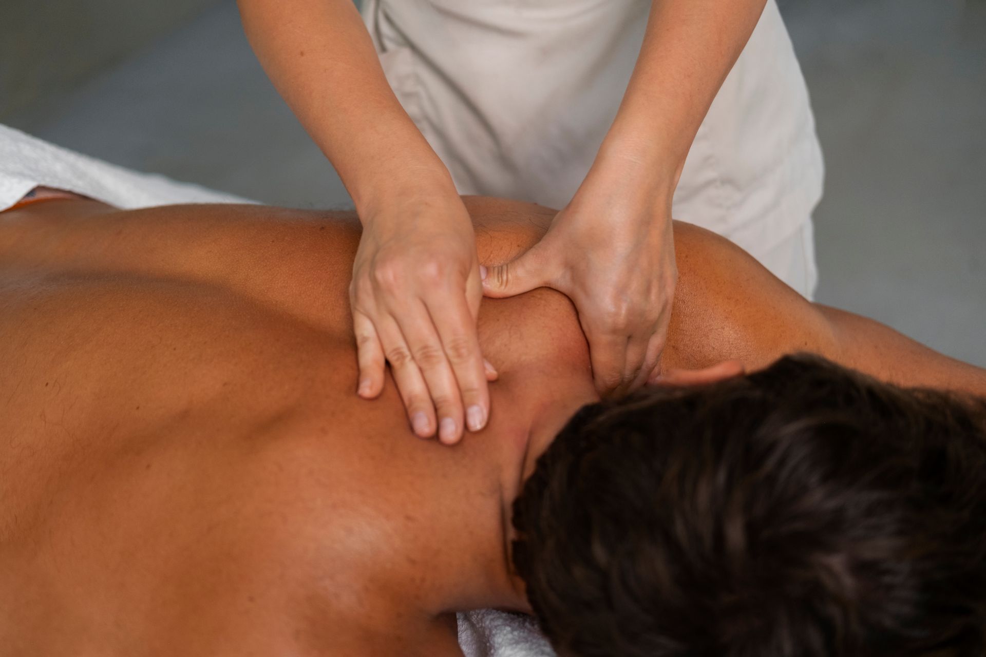 Hands massaging a person's shoulder and neck. Person lies face down on a massage table.