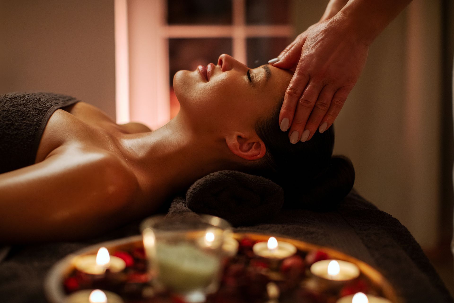 A person receives a head massage while lying on a spa bed, with glowing candles and petals in the foreground.