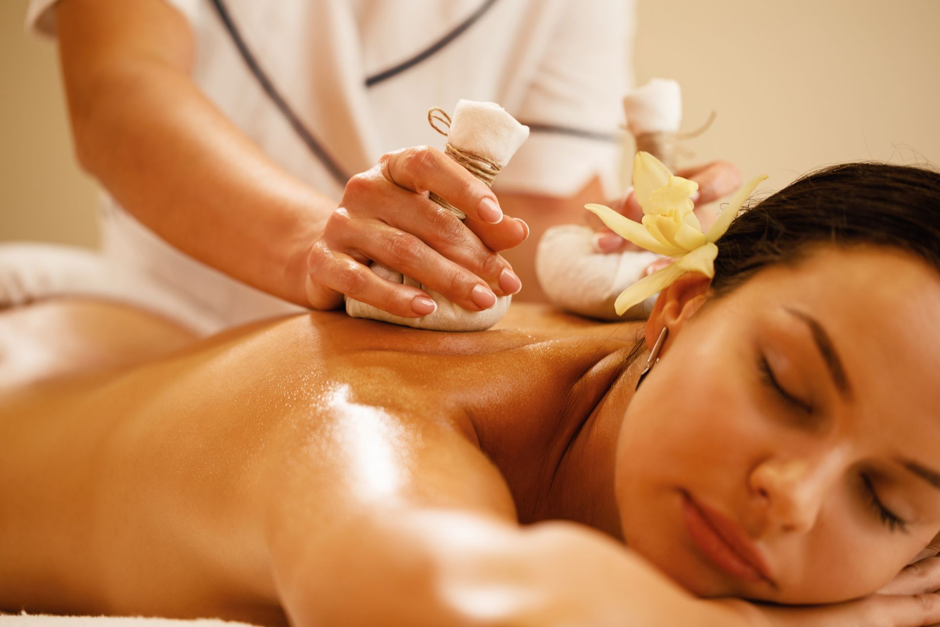 Woman receiving a back massage with herbal compress bags at a spa, with a flower in her hair.