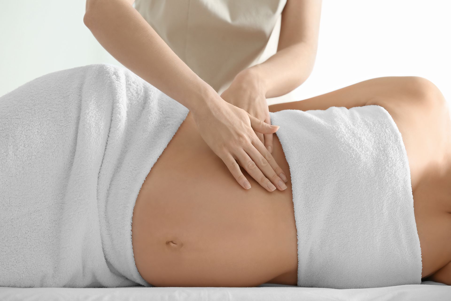 Pregnant person receiving belly massage, wrapped in a white towel, with a therapist's hands on the abdomen.