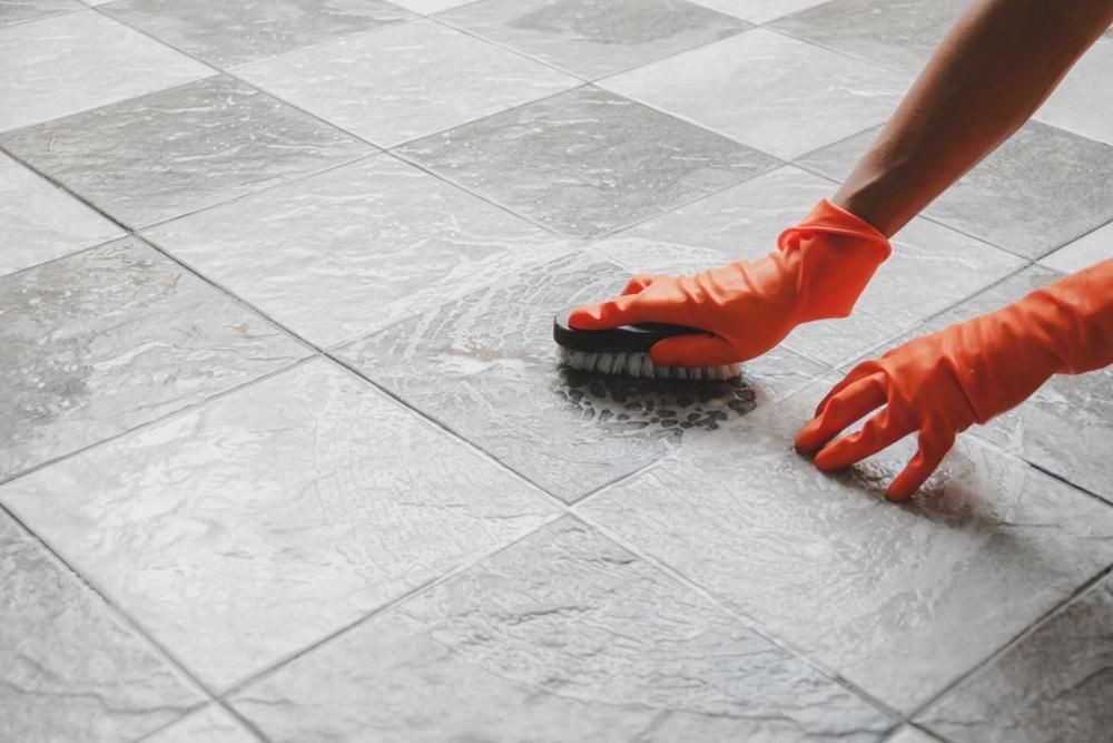 Hands in Orange Gloves Scrubbing a Gray Tile Floor With a Brush — AB QUICK Painting & Maintenance Services In Mareeba, QLD