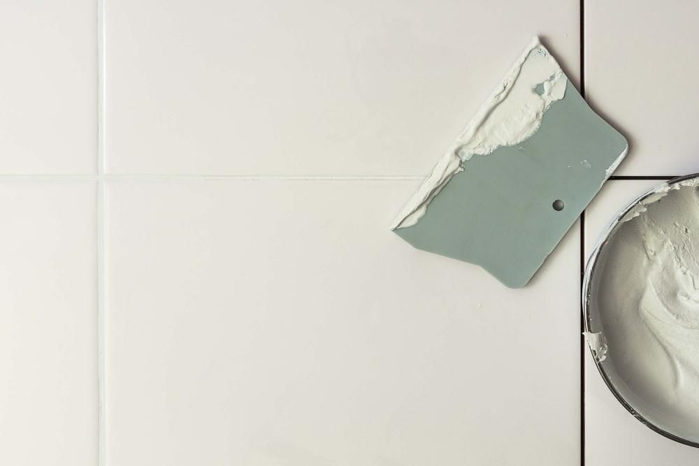 A Grout Trowel and Bowl of Grout on White Bathroom Tiles — AB QUICK Painting & Maintenance Services In Mareeba, QLD