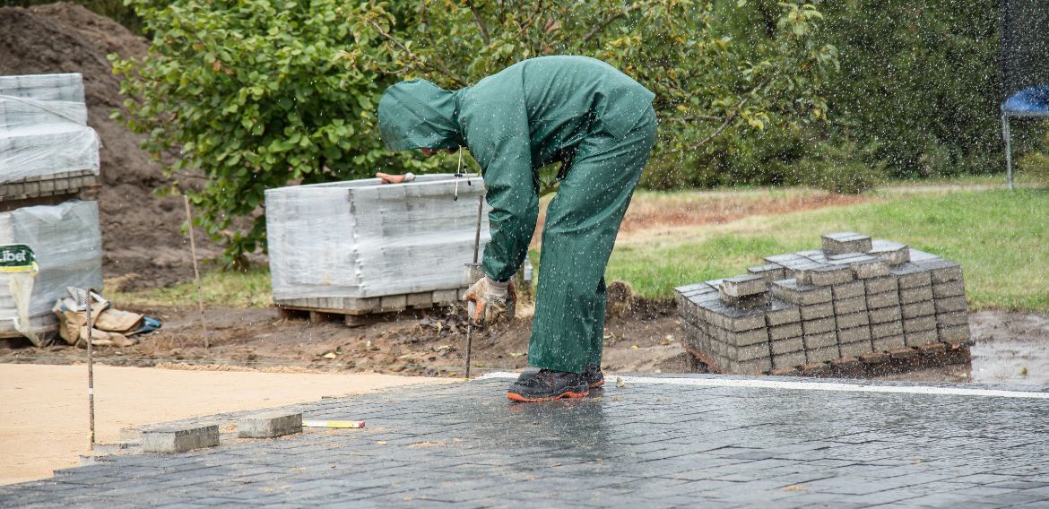 A person in green rain gear is working on laying pavers in the rain.