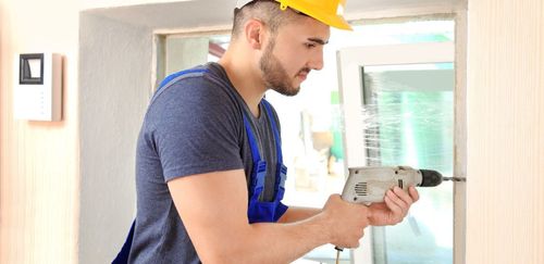 A construction worker drilling into a wall wearing a yellow hard hat and blue overalls.