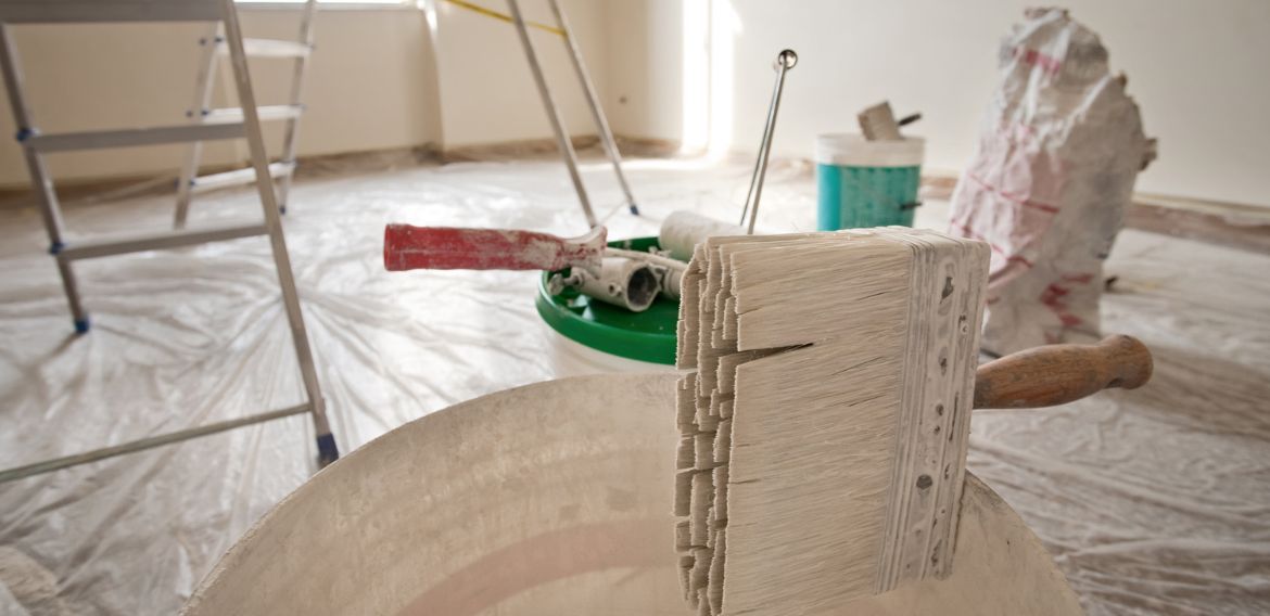 Room renovation in progress with paint supplies. Ladder, paint rollers, and a bucket on covered floor.