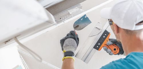 Person in blue shirt and white cap applying drywall compound with tools.