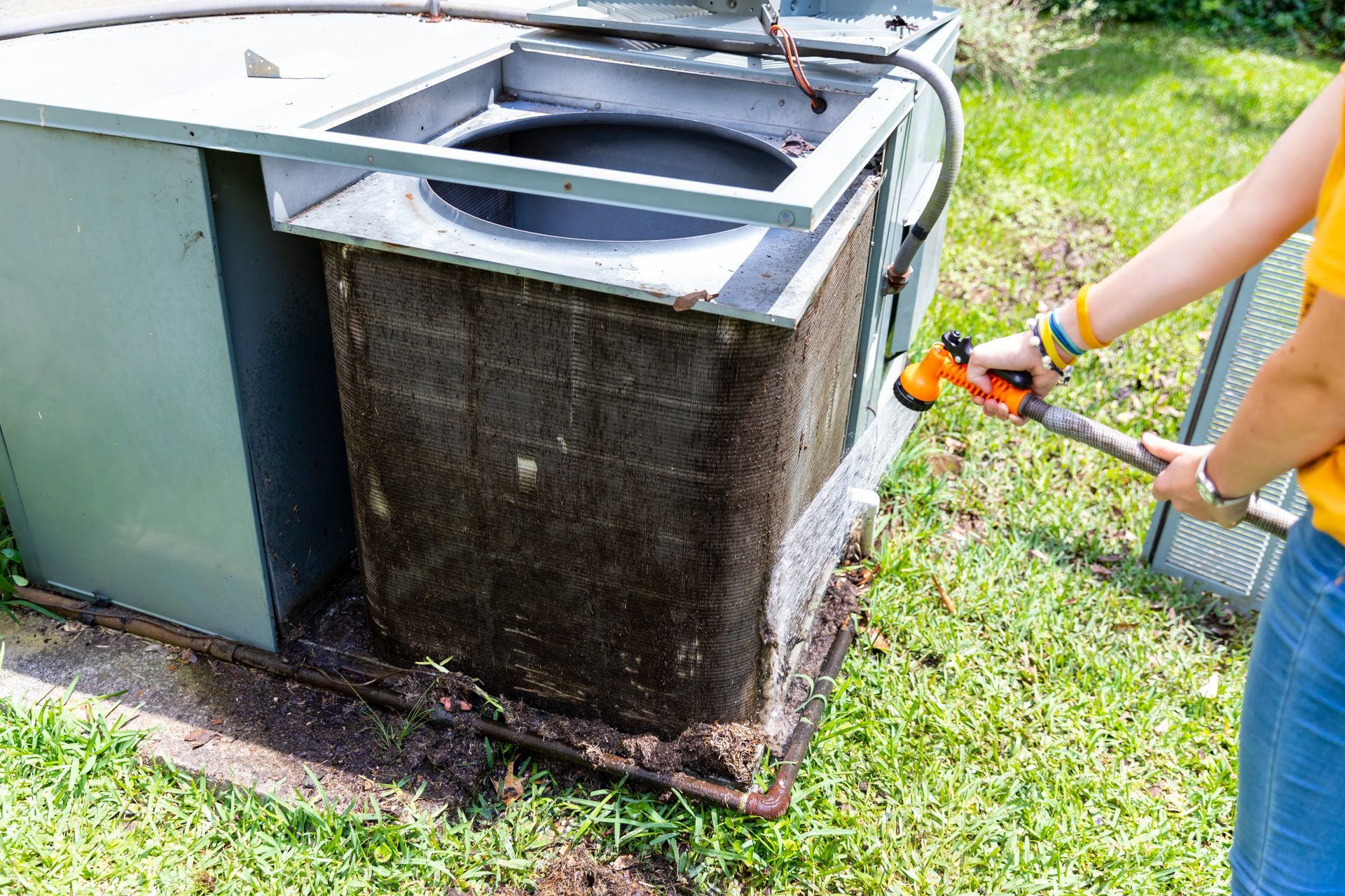 Person sprays outdoor AC unit with a hose, cleaning the coils on a grassy lawn.