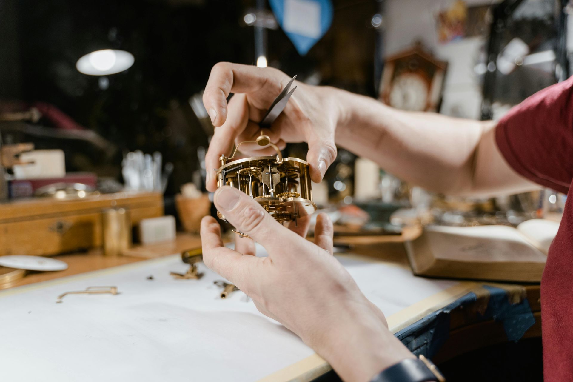 Man working on clock parts in a workshop, surrounded by antique clocks on the walls.