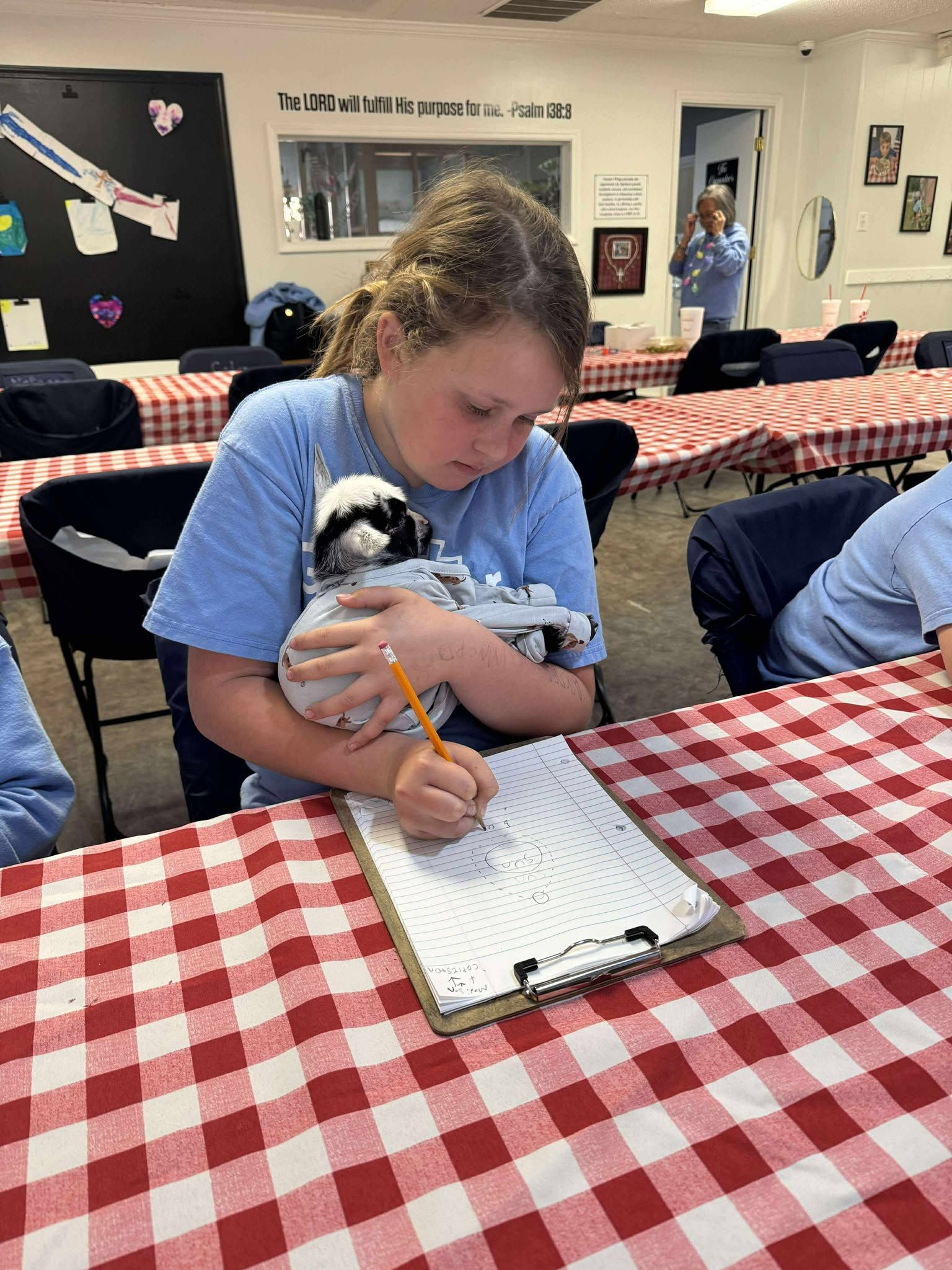 A girl is sitting at a table holding a puppy and writing on a clipboard.