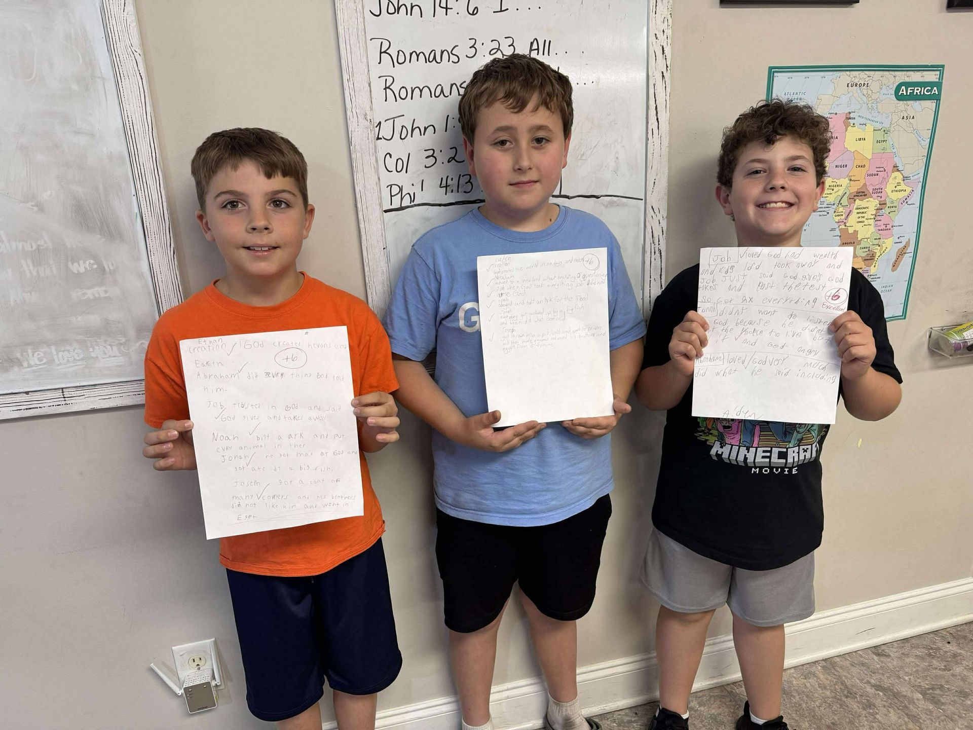 Three young boys are holding up papers in front of a whiteboard.