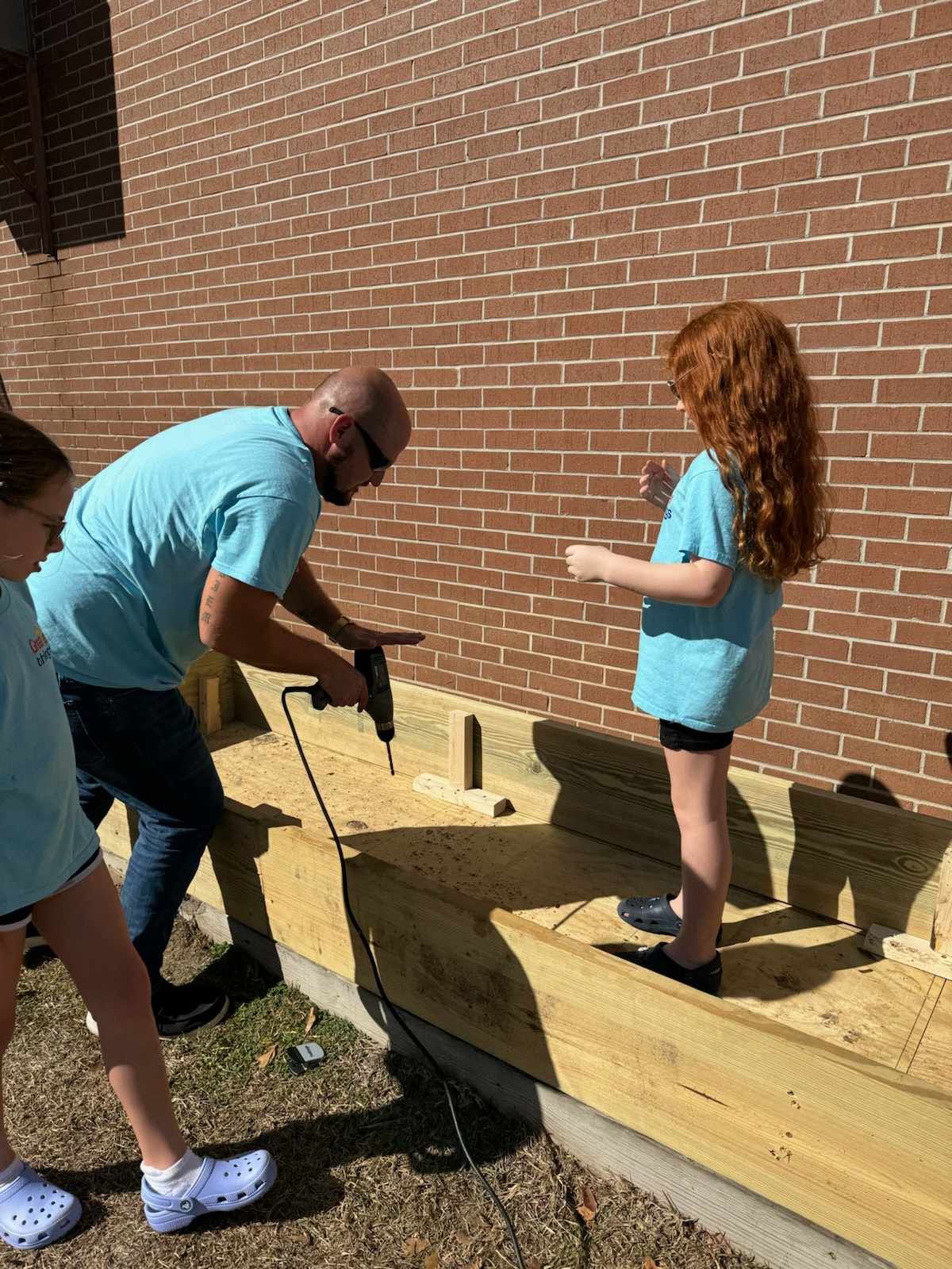 A man and a little girl are working on a wooden structure in front of a brick building.