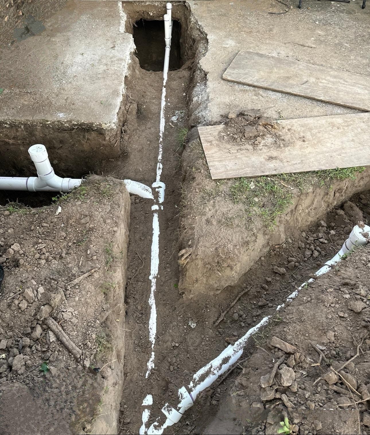 Open trench with white PVC pipes and fittings beside concrete slabs during utility work