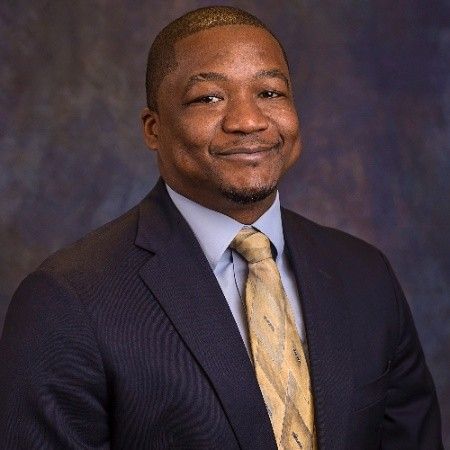A person in a dark suit, light blue dress shirt, and gold patterned tie, smiling against a dark blue mottled background.
