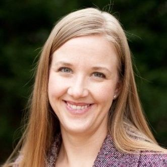 A person with long blonde hair and a friendly smile, wearing a textured purple blazer against a blurred green background.