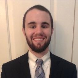 A person with a beard, wearing a black suit, light shirt, and patterned tie, smiling against a light-colored door.