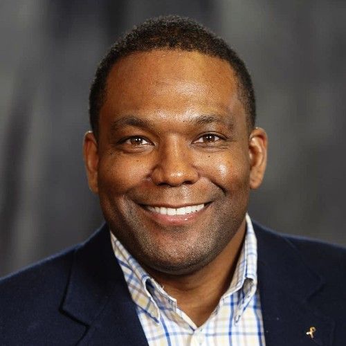 A man smiling, wearing a blue blazer and a plaid collared shirt against a grey background.