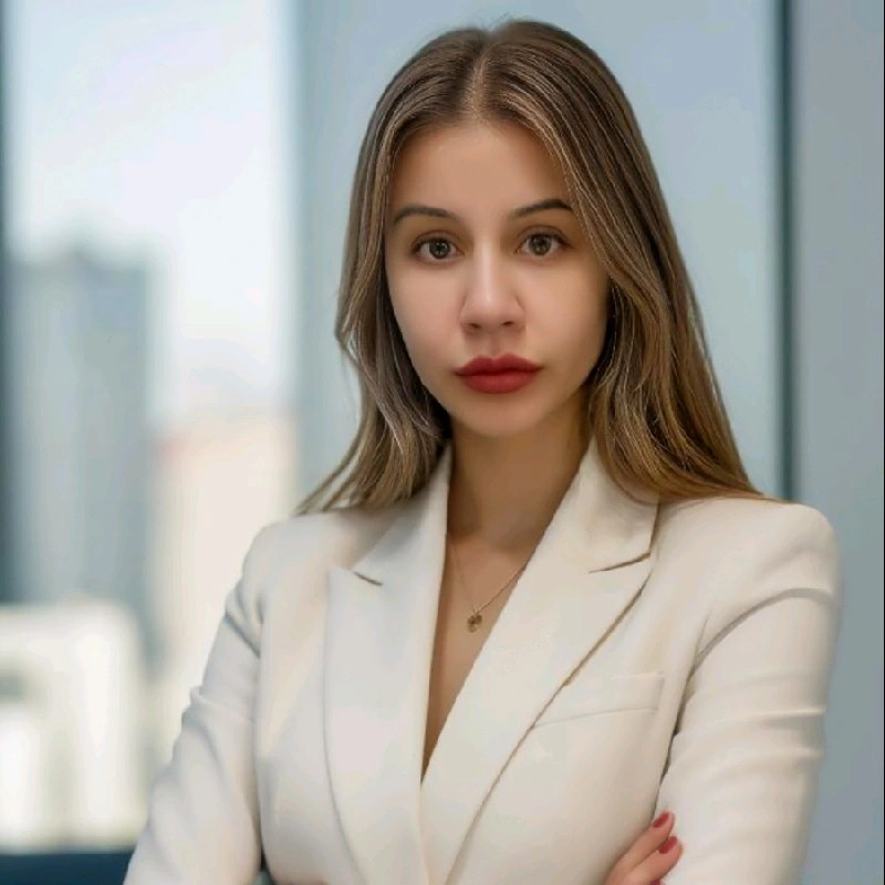 A person with shoulder-length hair wearing a cream blazer, posing with arms crossed in a brightly lit office setting.