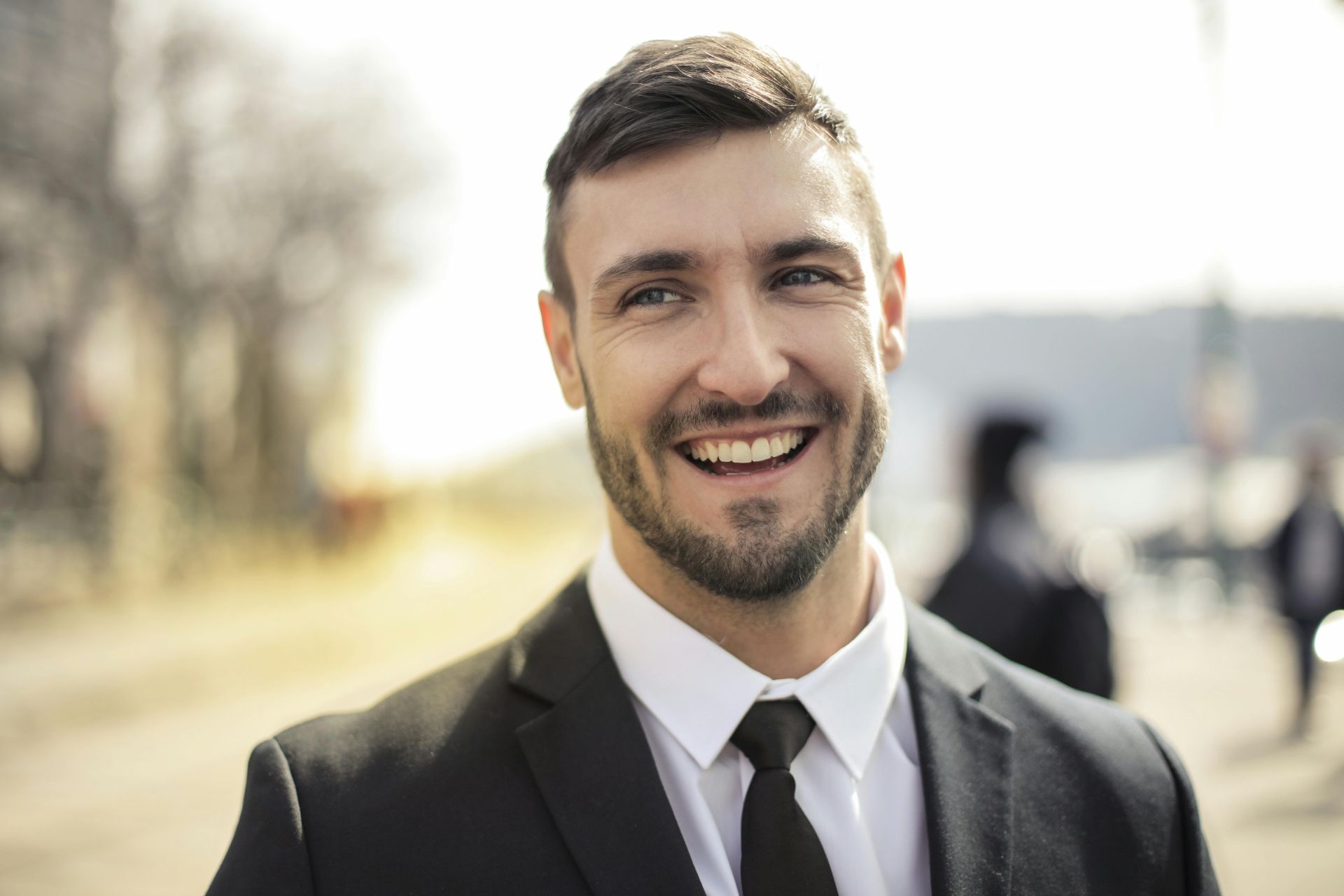 Smiling person with a beard, wearing a black suit and white shirt, outdoors with a softly blurred background.