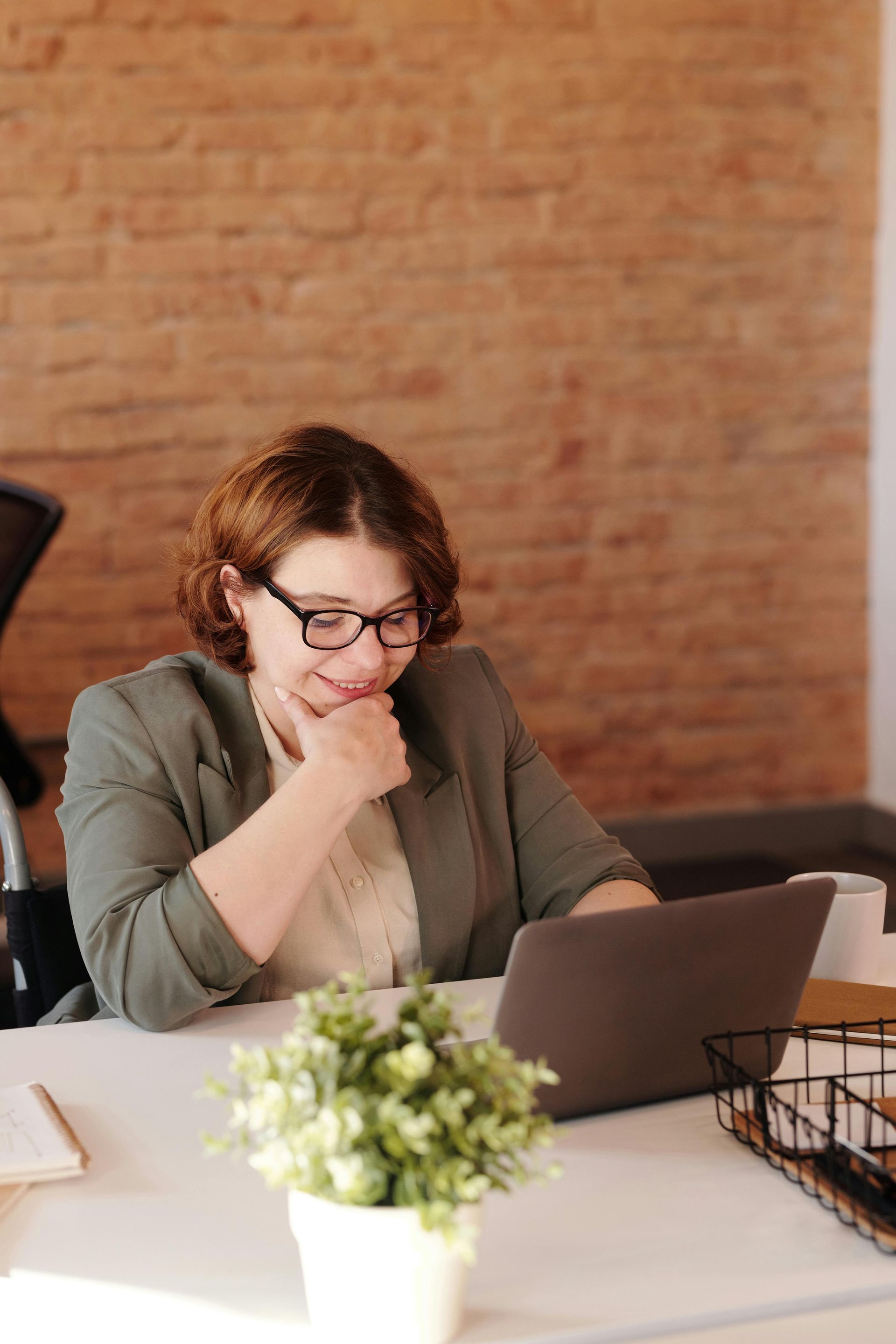 A person wearing glasses and a green blazer smiles while looking at a laptop on a desk in front of a brick wall.