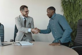 Two professionals in business attire shake hands over a desk in a bright office with a green wall.