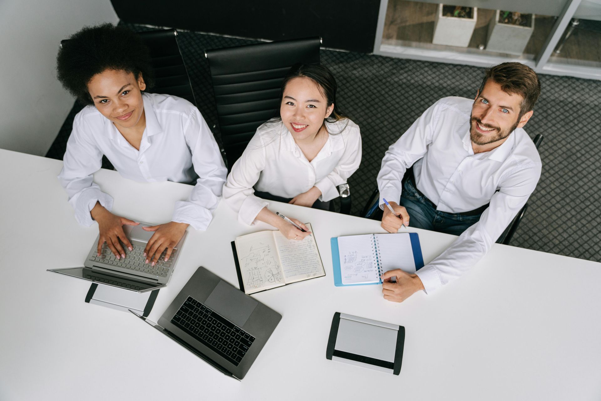 Three people sitting at a desk with laptops and notebooks, looking up at the camera in an office setting.
