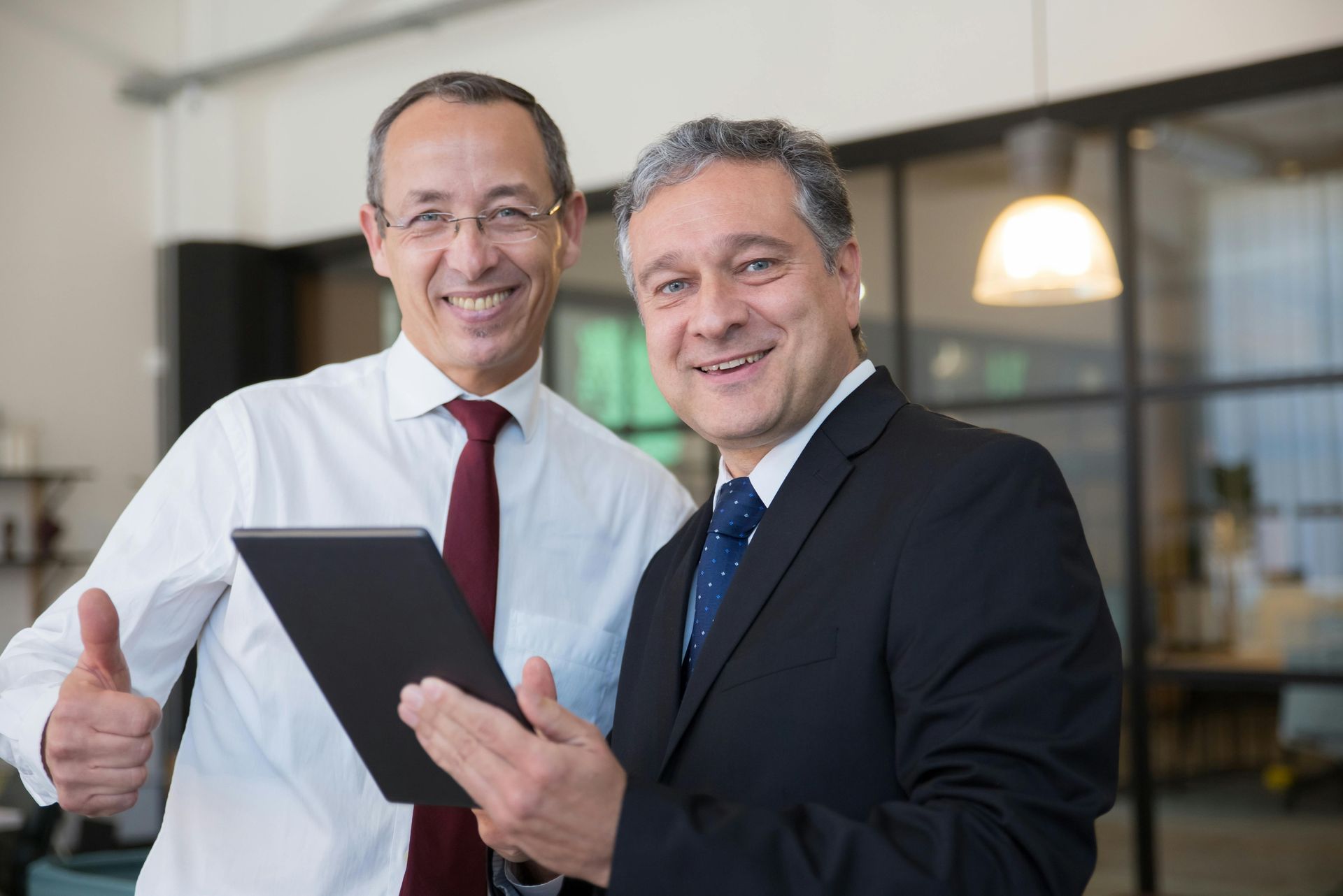 Two professionals in business attire stand in an office, smiling as they look at a tablet with one giving a thumbs up.