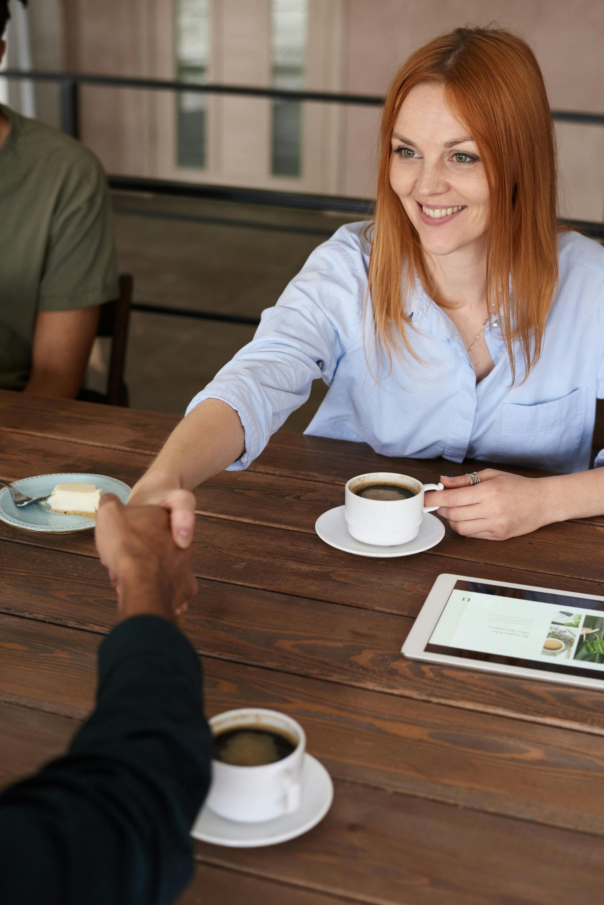A woman is shaking a man 's hand while sitting at a table with a cup of coffee.