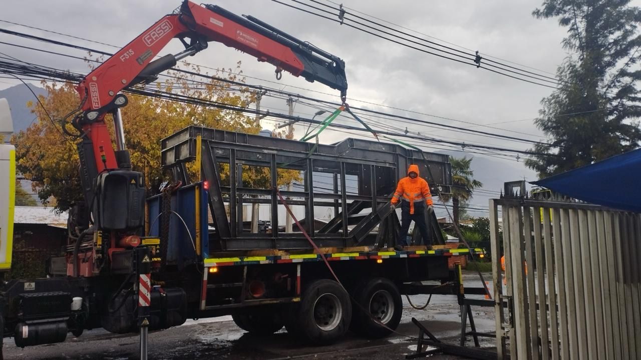 Un camión con grúa levanta una estructura metálica. Trabajador con chaleco naranja, día nublado.