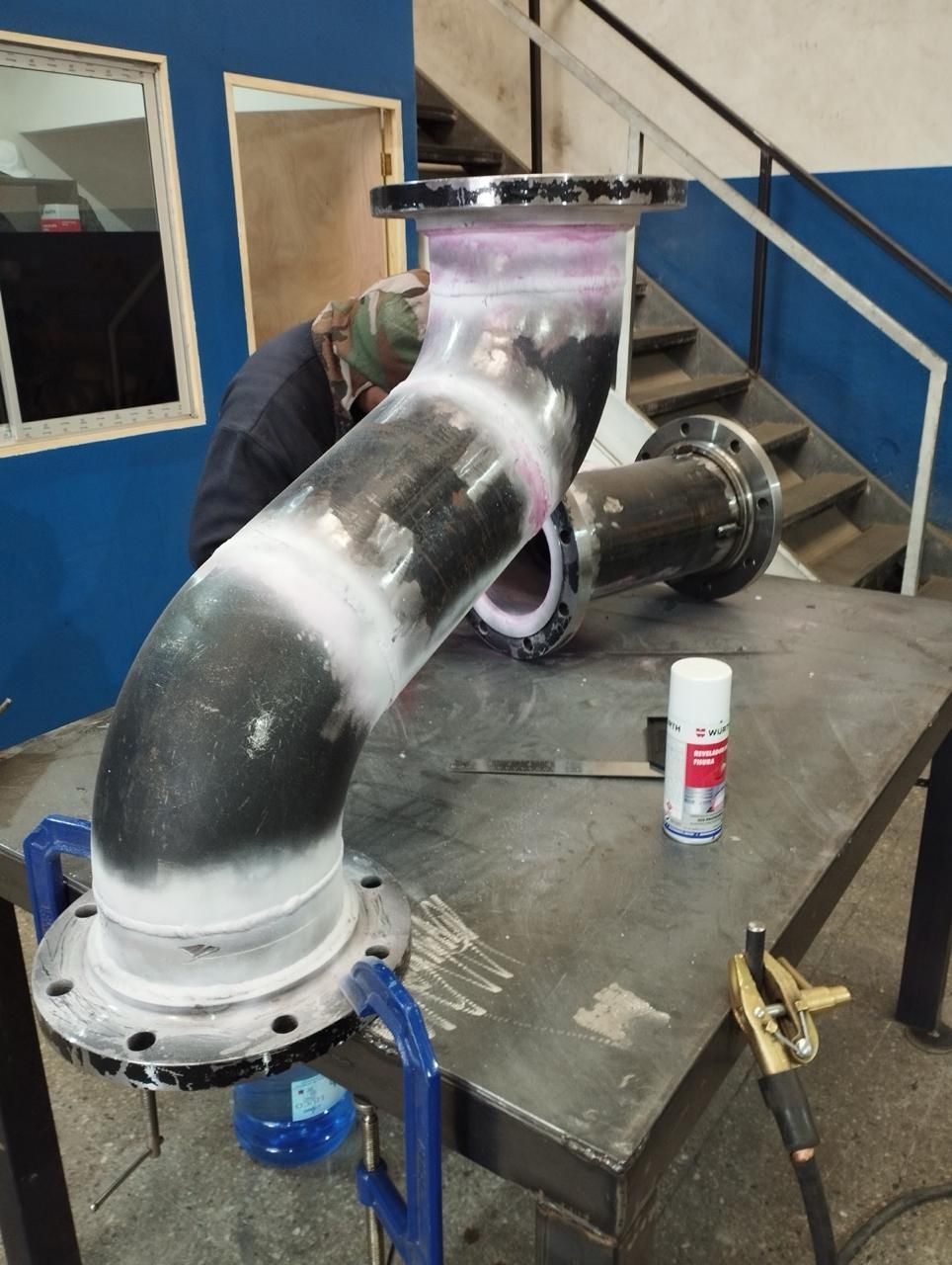 Soldador inspeccionando tubería con tinte rosa, banco de trabajo, entorno industrial.