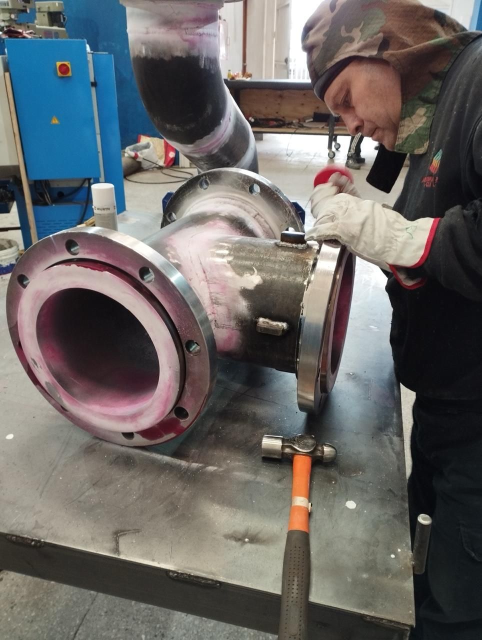 Un trabajador inspecciona un accesorio de tubería grande con un martillo. Se aplica tinte rosa al metal.
