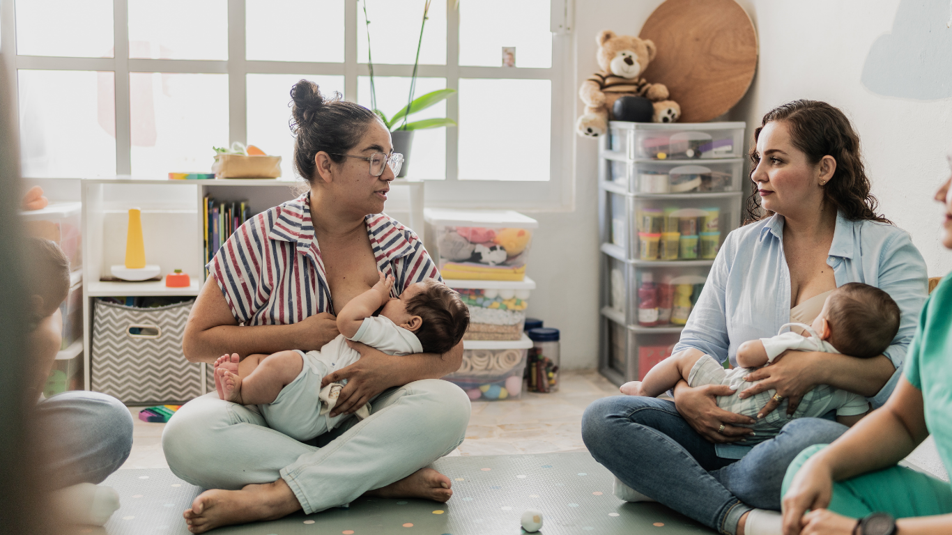 Two women seated on the floor holding babies, talking in a bright playroom.