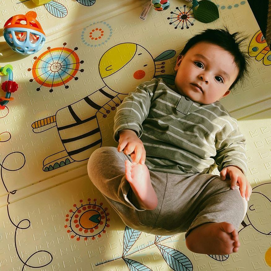 Baby lying on a patterned play mat, reaching toward a colorful toy.