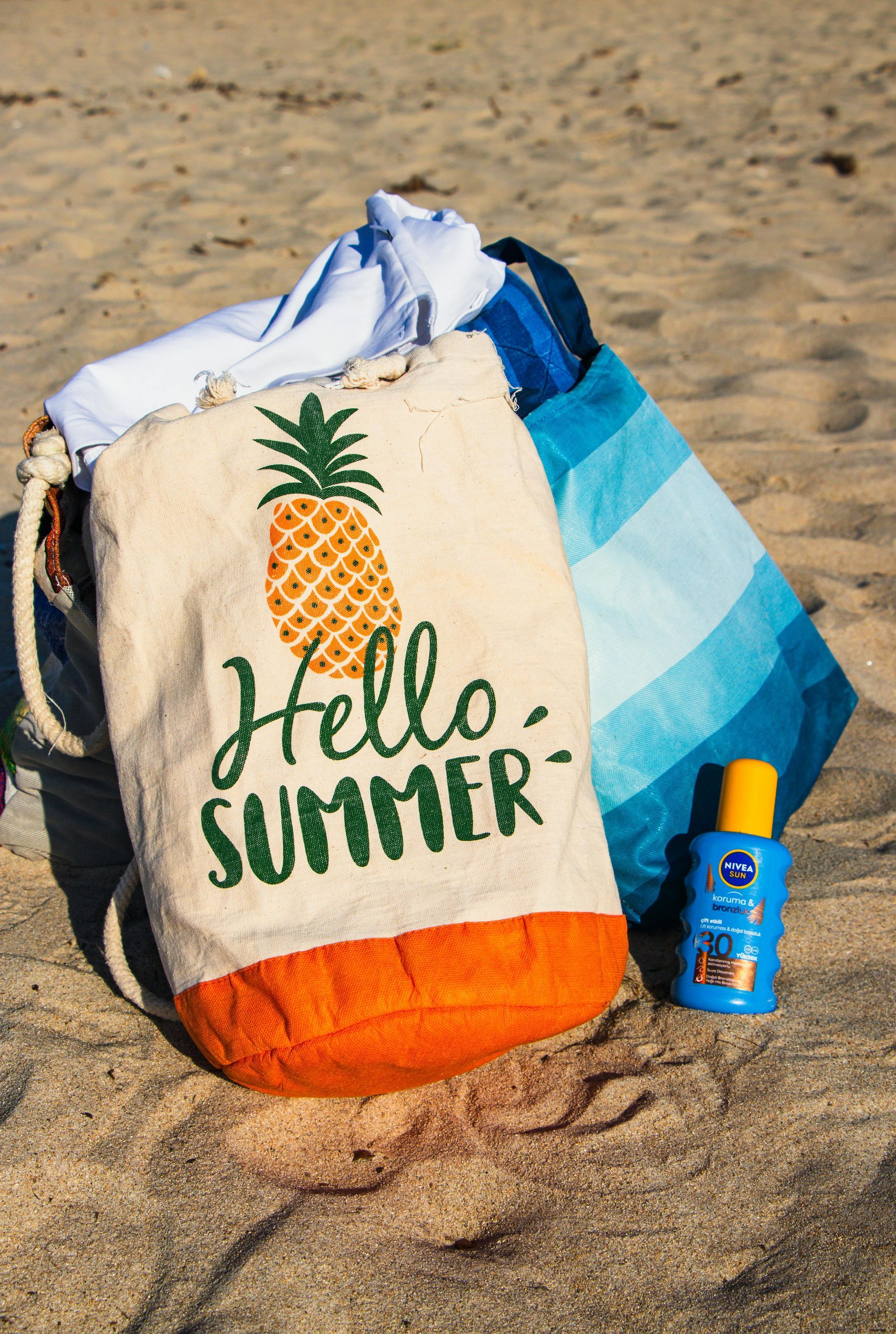 Beach bag with pineapple and “Hello Summer” text beside a sunscreen bottle on sand
