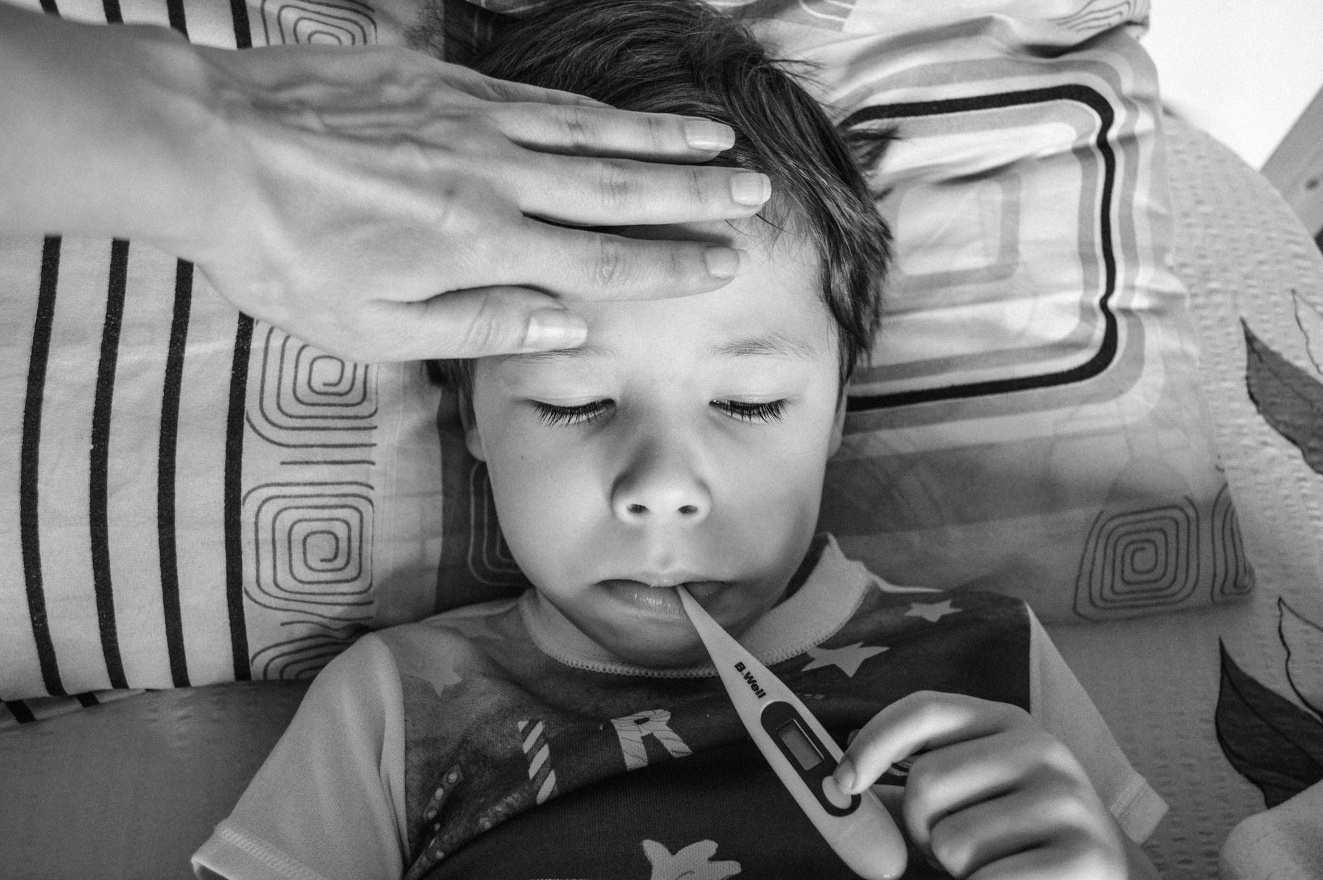 Child lying in bed with a thermometer, while an adult hand rests on their forehead.