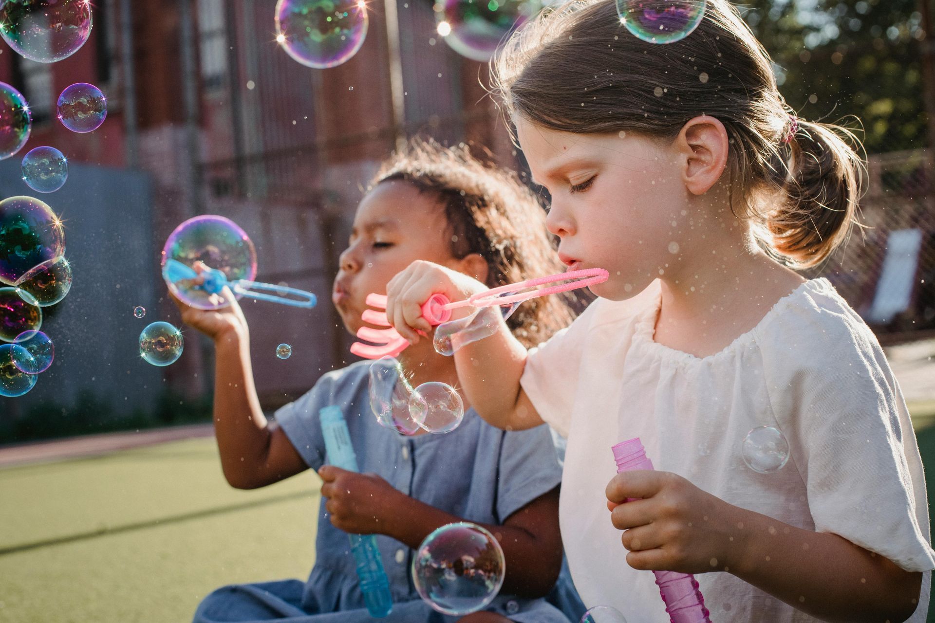 Two children blowing bubbles outdoors, with sunlight and a green playground in the background
