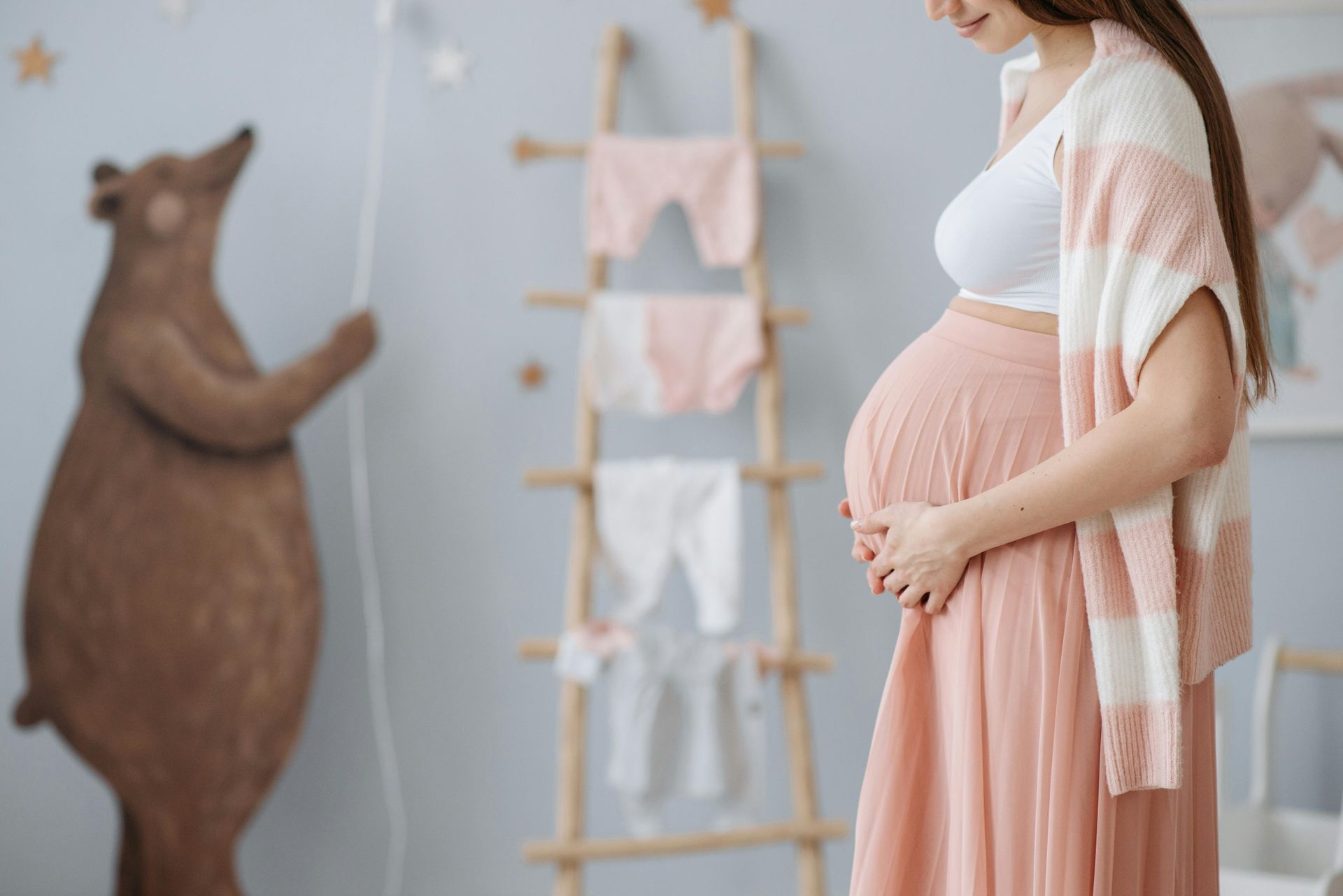 Pregnant woman in a pink dress and shawl standing beside a nursery shelf with baby clothes and a teddy bear