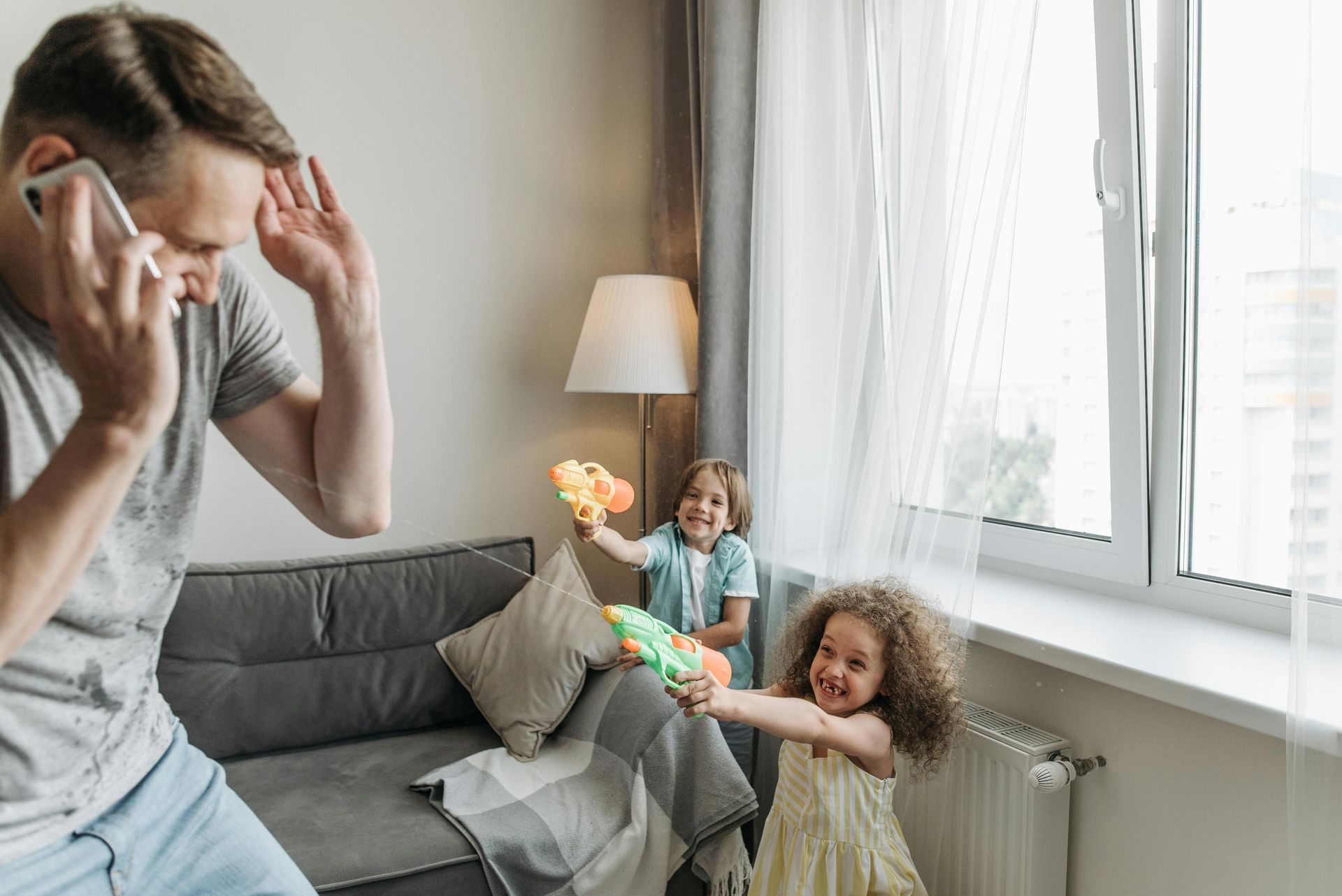 Man holding his head on a phone while two children play near a bright window in a living room