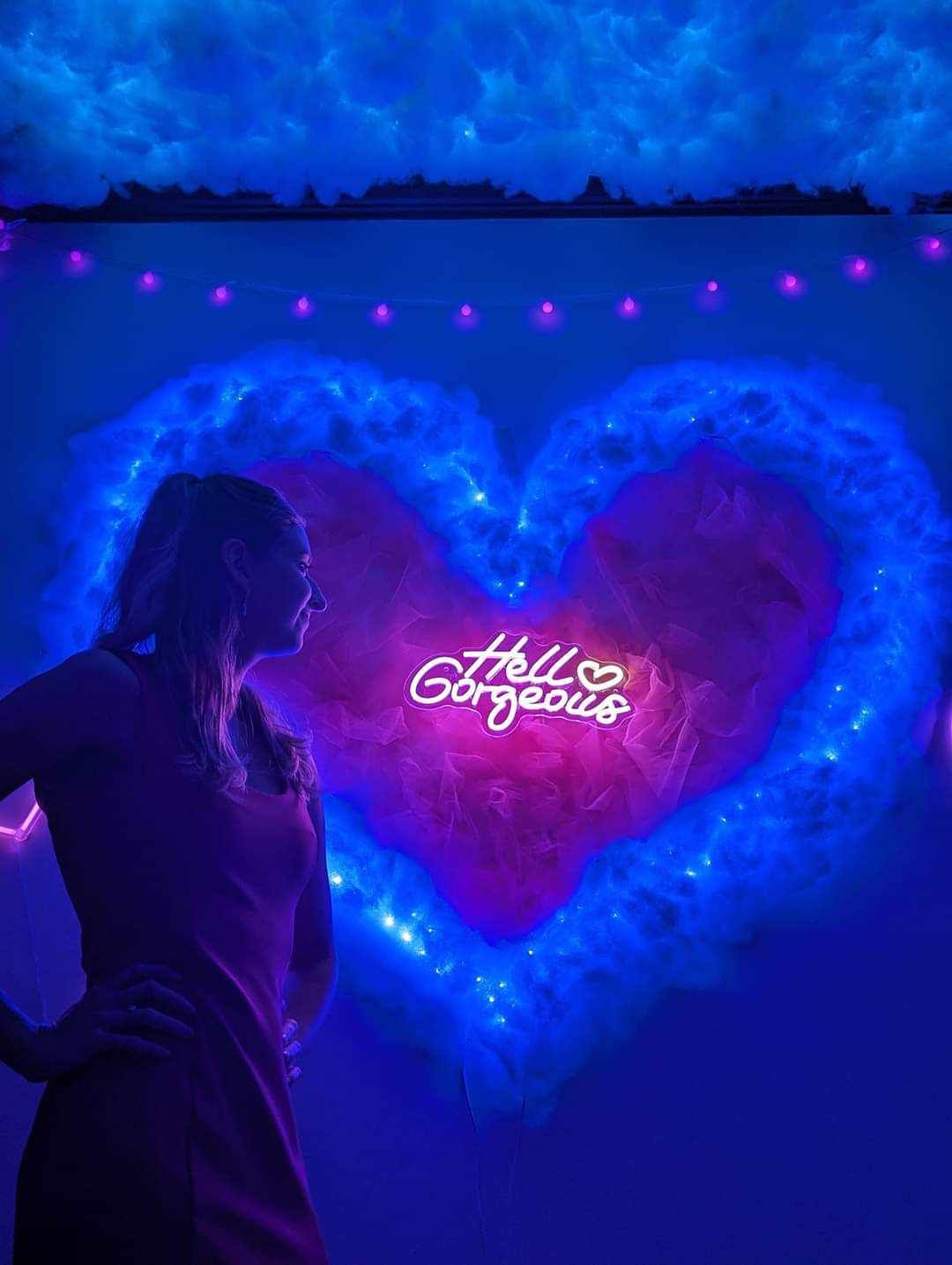 A woman is standing in front of a heart shaped neon sign that says `` hello gorgeous ''.