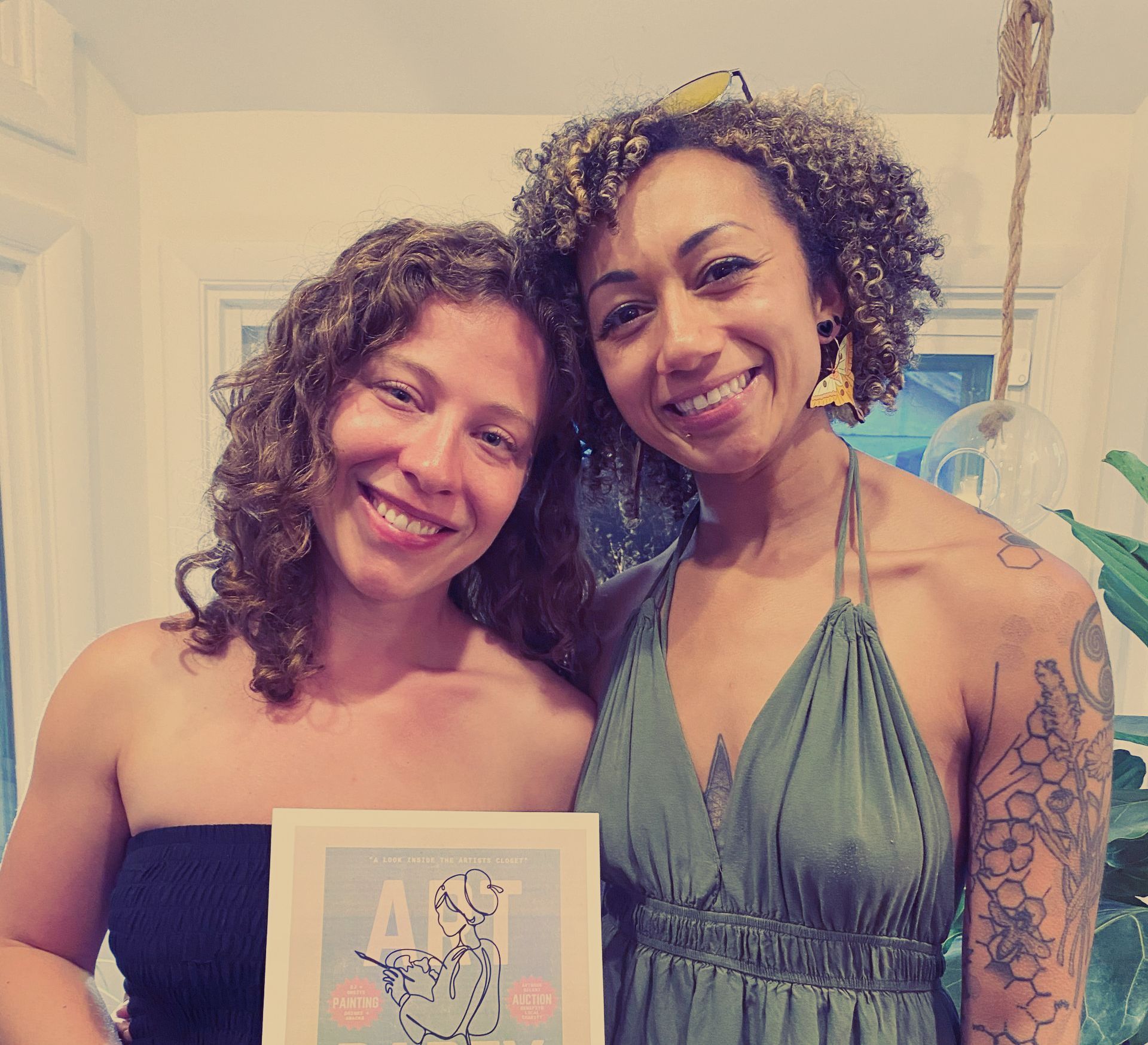 Two women are posing for a picture and one of them is holding a framed picture with the word art on it