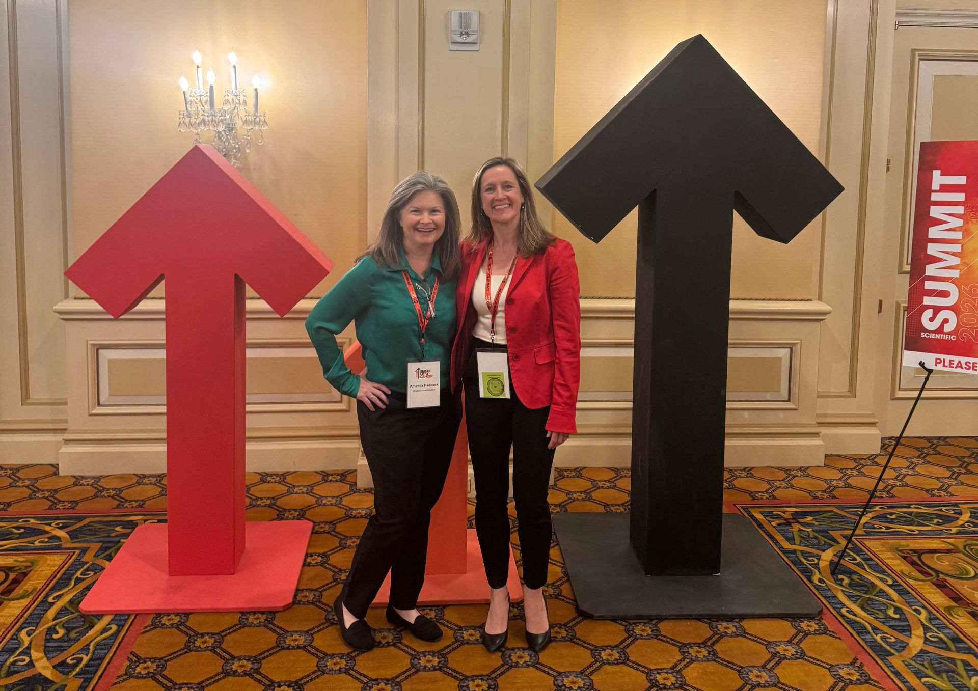 Two women pose with large red and black arrow cutouts at a summit.